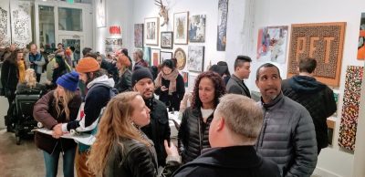 Interior view of an art gallery opening. Art is displayed on the walls salon style, and the space is full of visitors.