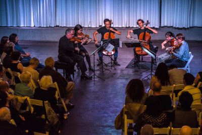 A group of string instrument players sit in a performance space, playing for a full audience.