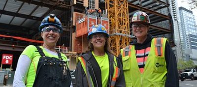 Three people wearing hard hats and safety vests stand in front of a construction site.