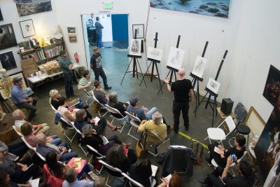 Art studio with a seated audience watching a light skinned masc person giving a lecture in front of five drawings on easels.