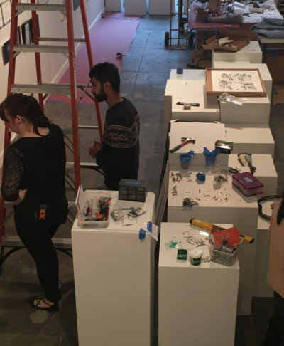 Aerial view of an art gallery in the midst of an installation. Two people stand thoughtfully in front a wall, next to a ladder.