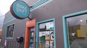 Outside of a grey building housing an arts venue, a circular blue sign above the orange door reading "Make Shift"
