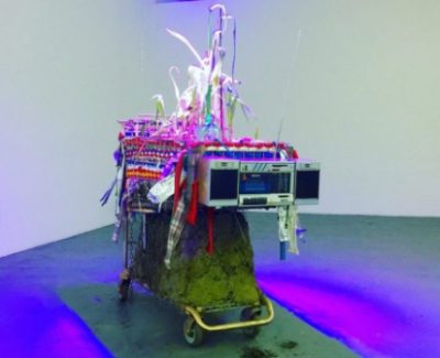 Brightly colored art sculpture consisting of a boom box on a decorated shopping cart installed in a white walled gallery.