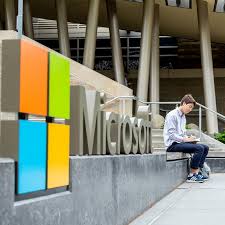 Light skinned person in business casual sits writing in a notebook on a step in front of an office building. A sign reading "Microsoft" is in the foreground.