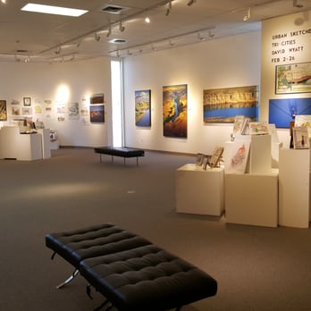 Interior view of a white walled art gallery. The walls have a variety of paintings hung, and there are two black benches in the middle of the space.