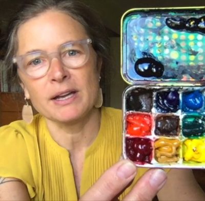 Femme person with translucent plastic rimmed glasses and a yellow shirt shows a used paint palette full of different paint colors.