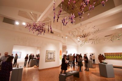 Interior of an art museum featuring white-walled exhibition spaces, 2D artwork hung on the walls and sculptural artwork installed in the center of the room. Crowds of viewers stand in small groups, looking at the work and chatting. A series of sculptural mobiles hang from the ceiling.