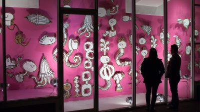 Exterior view at night of a storefront art installation featuring a pink painted wall, and cut out black and white floating drawings on board. Two silhouetted figures stand in front of the lit windows.