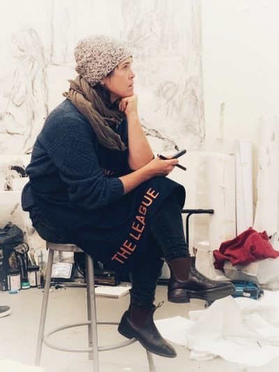 Person in a knitted beanie sits on a tall metal stool in a drawing studio, their chin resting on their hand thoughtfully. They are wearing an apron that reads "the league"