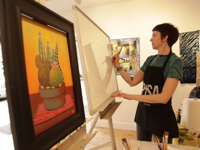 Person wearing a black work apron stands in an art studio in front of a white board, poised to make a mark.