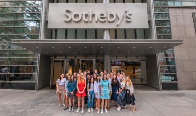 Group of young adults of varying ages, gender identities, and skin tones stand in front of a building entrance. Large grey sign above entrance reads "Sotheby's."