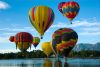A group of 8 hot air balloons flying over a lake, a small mountain and forest in the background.