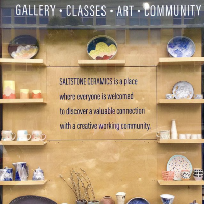 Wooden shelf display in a storefront setting, featuring several pieces of handmade ceramic artwork, including various mugs, cups, plates, and bowls. Text across the top reads "Gallery; Glasses; Art; Community." Central text reads "SALTSTONE CERAMICS is a place where everyone is welcomed to discover a valuable connection with a creative working community."