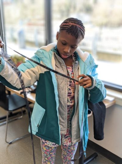 A dark skinned youth in a teal and blue winter coat looks over an outstretched roll of analogue photography film in a classroom setting.