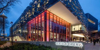 Exterior shot of a performing arts venue building, lit up at dusk. Small groups of people congregate both inside and outside the building. Lit sign on side of the building reads "Seattle Opera."