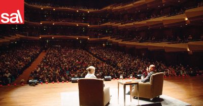A large multi-tiered indoor auditorium setting, full of people, showing two people on a stage from behind. They are both seated in brown armchairs, and are engaged in conversation. Between them is a small side table with two glass of water.