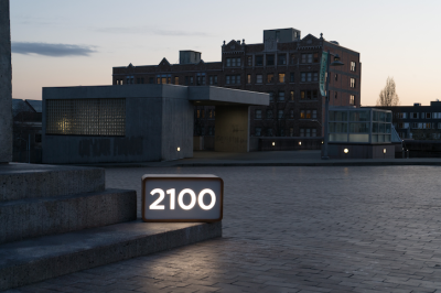 Distant view of a multi-story brown brick building at dusk. The foreground is focused on a brick paved street, with three steps on the left side of the frame. On the bottom step is a small wooden box, with glowing numbers that read "2100"