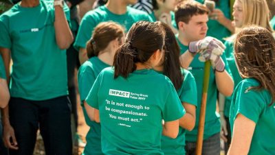 Group of students wearing matching green shirts in a work setting, one holding a shovel. Figure in the foreground is facing away from the camera, and their shirt reads "Impact; Spring Break 2017; 'It is not enough to be compassionate. You must act.' - Dalai Lama."