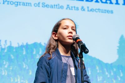 Youth in a blue jacket standing in front of a microphone, with a blurry banner in the background that reads "Program of Seattle Arts & Lectures"