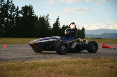 A small race car idles on pavement, set against a forested landscape, blurred mountains and clouds visible in the background.