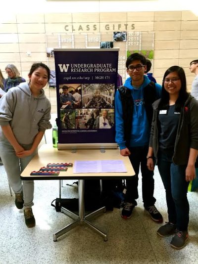 A group of three students stand in front of a tabling set up, which features flyers and a stand up poster advertising the Undergraduate Research Program.