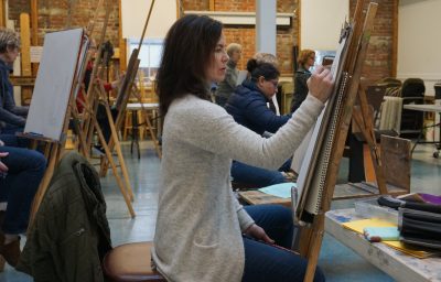 A group of adults in an art studio drawing on pads resting on individual easels.