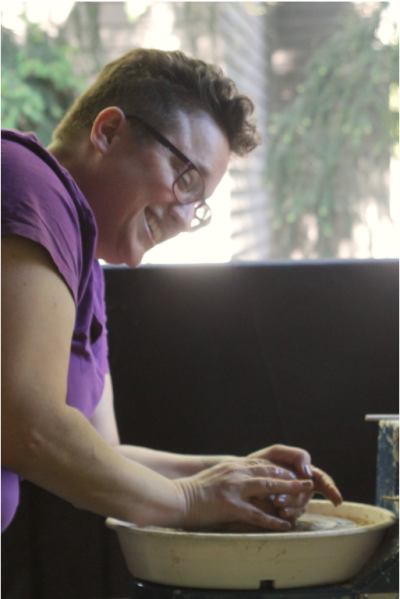 Person with short hair and glasses, wearing a purple tshirt, smiles while molding clay on a throwing wheel