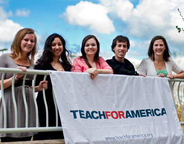 A group of five people stand in front of a white railing, holding a white banner that reads "Teach For America; www.teachforamerica.org".