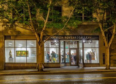 Exterior view of gallery windows at night, a crowd of people inside looking at art and mingling. Sign above the entrance reads "Patricia Rozvar Gallery."