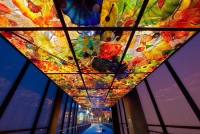 Hallway art installation featuring hundreds of multi-colored glass pieces behind a glass ceiling, lit from behind.