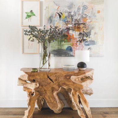 Hand made raw wood end table with a vase of flowers, lamp and ceramic sculptures. Three framed art pieces hang on the wall behind.
