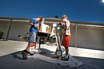 Two youths work outside on a wood sculpture using hand tools