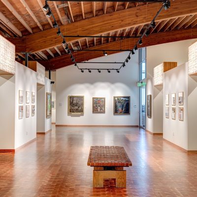 brightly lit museum gallery featuring painting and 2D work on the walls. In the center of the room is a wood bench.