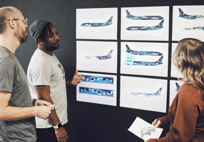 Three people view and discuss a wall full of design drawings featuring airplane spaces