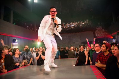 Person in a white suit performs onstage in a musical theater setting which an audience applauds
