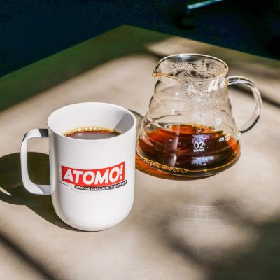 White coffee cup with a red logo reading "Atomo!" full of coffee sits next to a glass coffee pour over vessel.