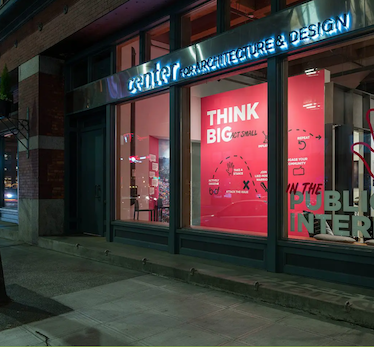 Lit storefront windows showing an education display with the words "Think Big" above infographics on red paper. A lit sign above reads "center for architecture & design"