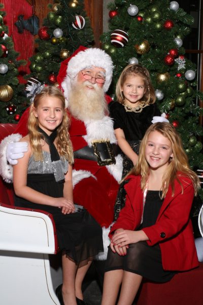Three young children dressed in holiday clothes gather around a man dressed as Santa Claus in front of a Christmas tree.