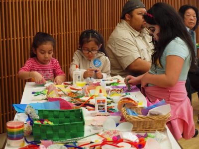 Group of three children gathered around a crafts table making artwork. One has face paint on.