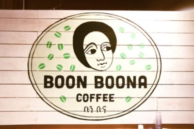 Circular logo painted on a white wood wall featuring a line drawing of a face, with green coffee bean imprints surrounding it. Black text reads "Boon Boona Coffee"