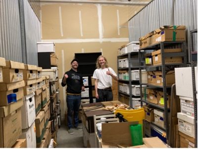Two people standing holding thumbs up signs in a full storage room, with shelves and boxes.