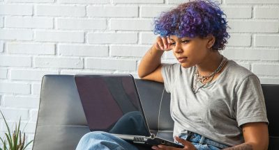 Light brown skinned person with dyed purple curly hair sitting in front of a laptop looking at the screen