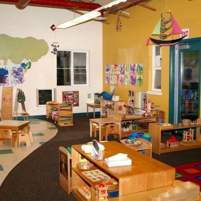 Brightly lit preschool classroom with play tables and toys neatly put away.