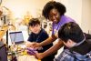 Person leaning over to help two young students in front of a computer station