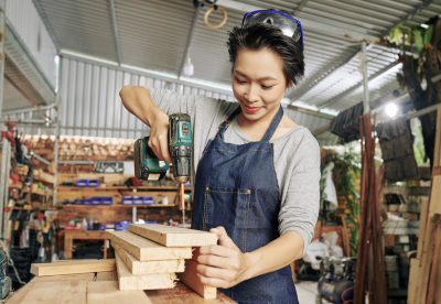Femme person with short hair wearing a shop apron uses a power drill on a stack of wood boards.