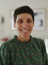 Headshot of Dr. Noa Brazg with short dark hair wearing a green patterned shirt and earrings, photographed indoors against a softly blurred background.