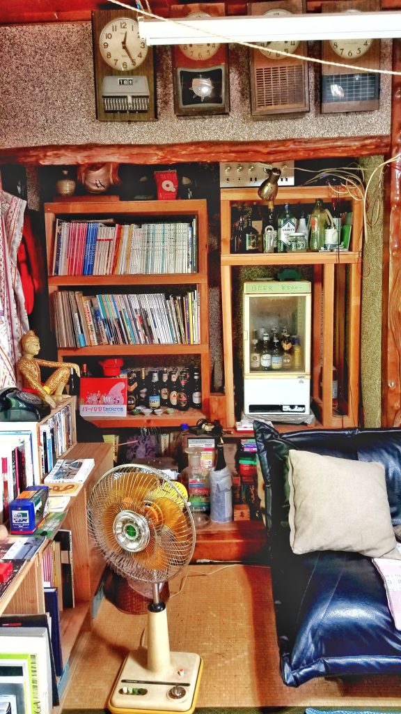 A corner of a room with bookshelves, clocks, a fan, and a sofa