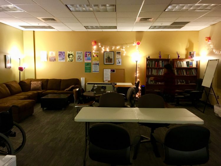A wide view of the D Center in MGH 024, warmly lit with posters and fairy lights strung on the wall. To left is a brown sectional couch, in the center are meeting tables with chairs, and to the right are two bookcases filled with books and games.