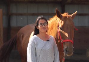 Grace standing in a light blue sweater with a microphone on her next to a brown quarter horse with a red halter on