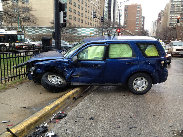 A blue car, heavily damaged on the front right and driven onto the sidewalk. Yellow side curtain air bags are deployed. Photo by Steven Vance.